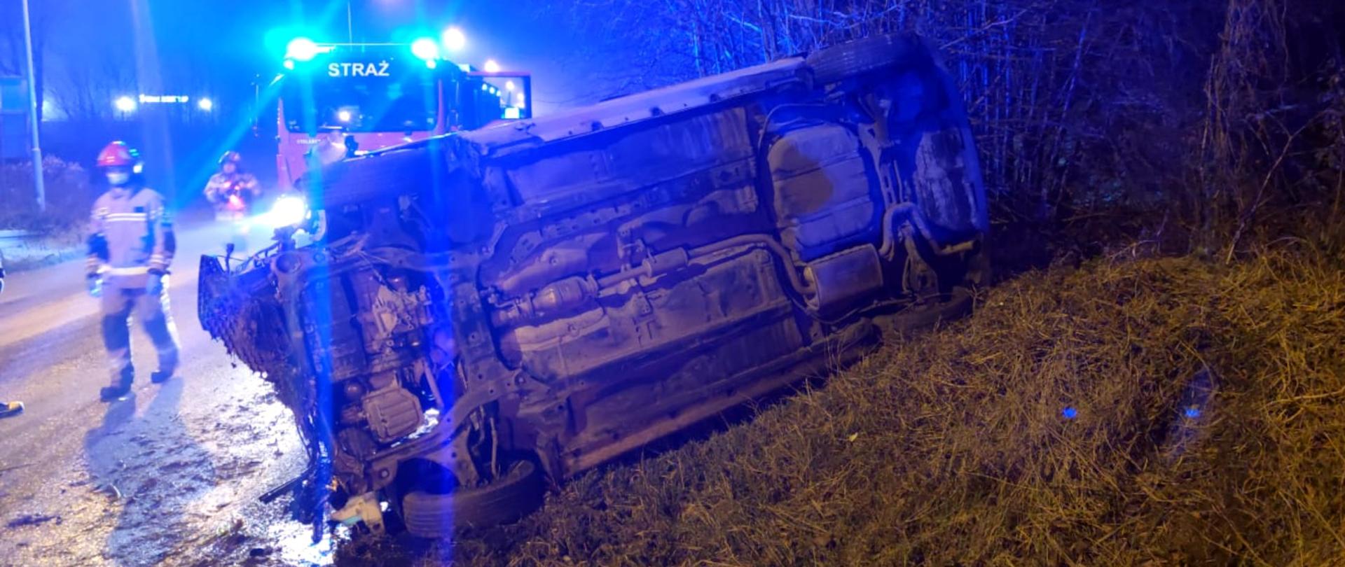 Zdjęcie przedstawia na pierwszym planie przewrócony samochód osobowy, obok pracujących strażaków, w tyle samochód ratowniczo - gaśniczy z włączonymi światłami ostrzegawczymi.