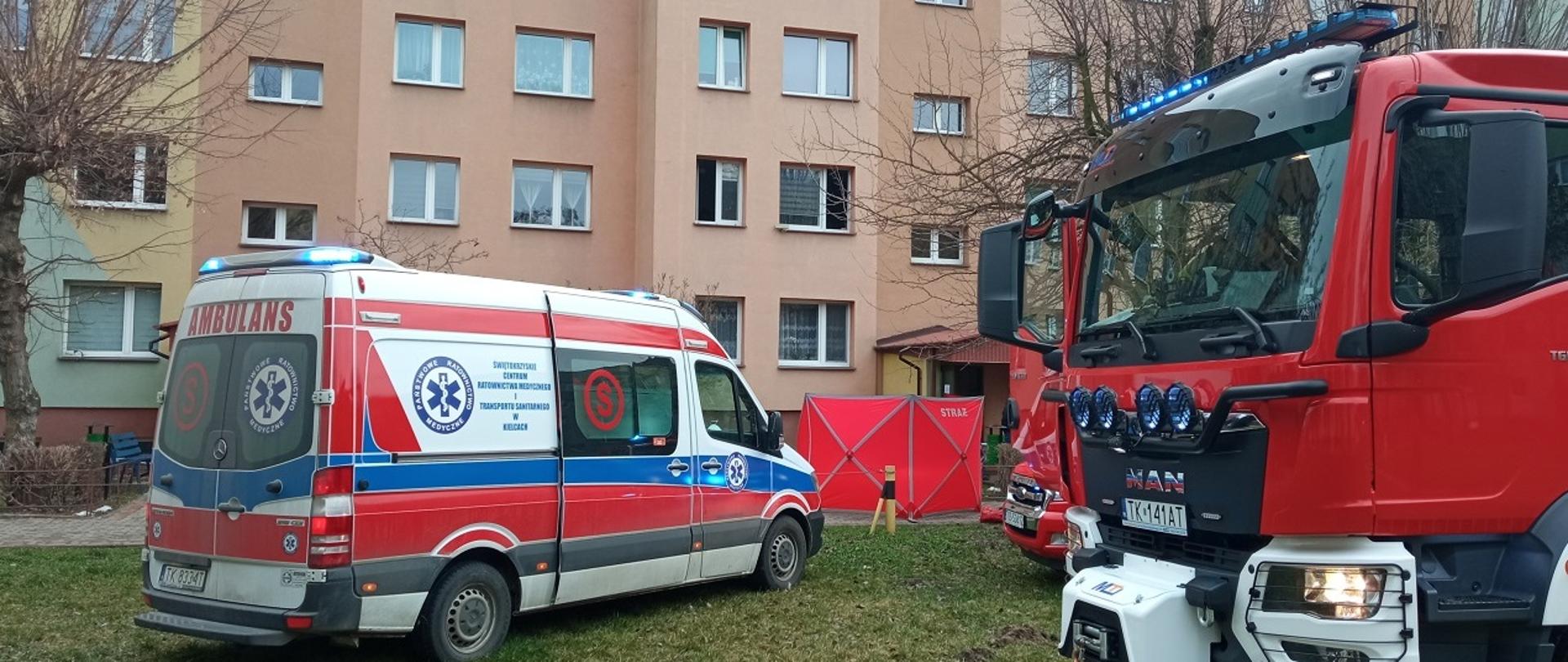 Na zdjęciu przed budynkiem mieszkalnym wielorodzinnym widać karetkę pogotowia oraz parawan zasłaniający osobę poszkodowaną. Obok stoi wóz strażacki. Zdjęcie wykonane w aurze pochmurnej w porze nocnej. 