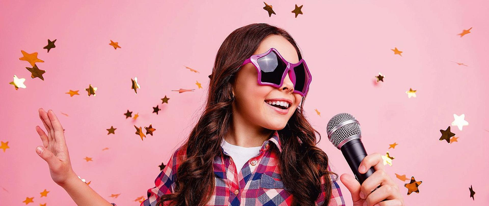 Plakat informujący o Jarocińskich Warsztatach Wokalnych 2023. W centralnym punkcie plakatu znajduje się zdjęcie dziewczynki w okularach w kształcie gwiazdy, śpiewającą do mikrofonu. Zdjęcie posiada różowe tło.