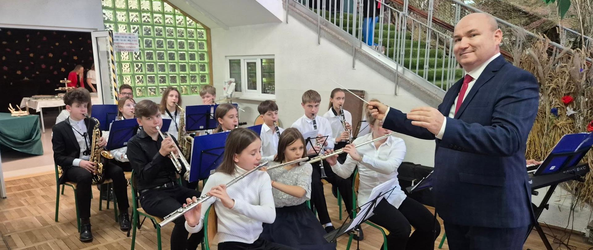W kadrze uczniowie orkiestry dętej podczas gry w pozycji siedzącej. Po prawej stronie pozuje do zdjęcia uśmiechnięty dyrygent - Tomasz Winczewski.