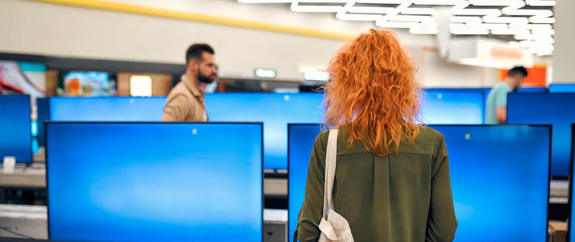 Zdjęcie przedstawia młodą kobietę w zielonej koszuli stojącą wewnątrz sklepu ze sprzętem RTV. W tle widać innych klientów oraz kilka nowoczesnych telewizorów. 