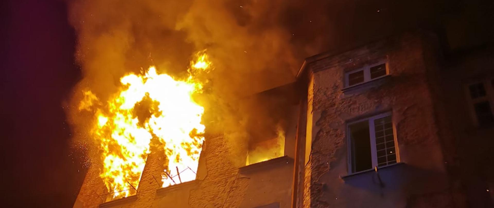 Zdjęcie w porze nocnej. Widać pomieszczenia objęte pożarem. Ściana kamienicy oświetlona sztucznym światłem. Występuje zadymienie.