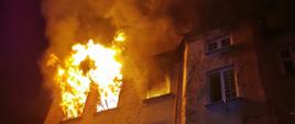 Zdjęcie w porze nocnej. Widać pomieszczenia objęte pożarem. Ściana kamienicy oświetlona sztucznym światłem. Występuje zadymienie.