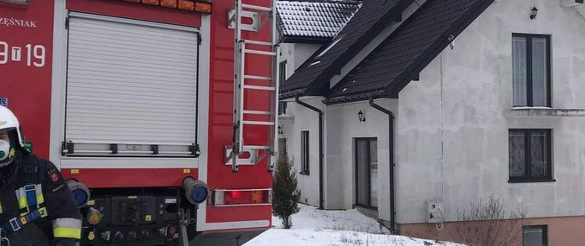 Zdjęcie przedstawia samochód strażacki na tle domu jednorodzinnego, w którym doszło do zdarzenia. Nie widać dymu ani ognia.