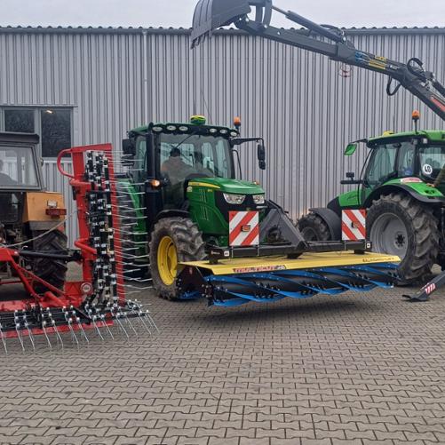 Trzy ciągniki rolnicze z podczepionym osprzętem przed halą z szarej blachy