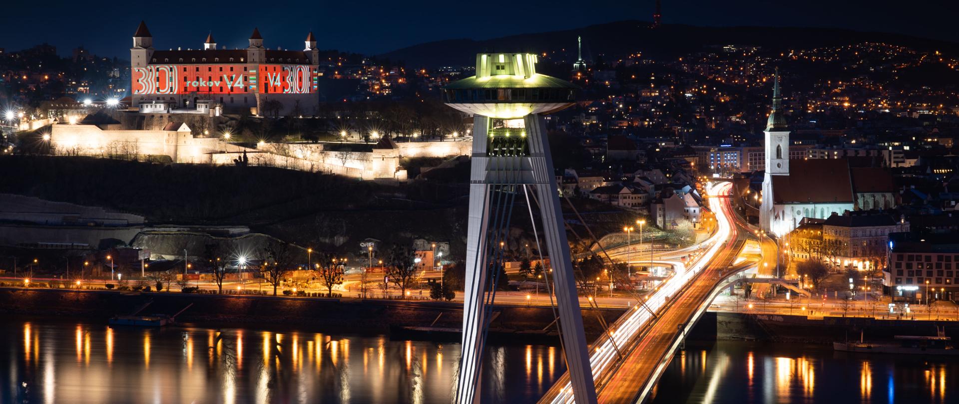 Nocne ujęcie starówki Bratysławy z podświetlonym Zamkiem Bratysławskim z logo z okazji 30-lecia powstania Grupy Wyszehradzkiej