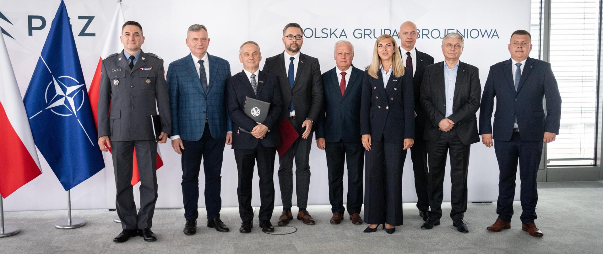 Family photo. Grupa osób na tle ścianki z logotypem PGZ. Z lewej strony flagi - Polski i NATO.