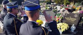 Widok z tyłu. Trzech strażaków PSP salutuje na cmentarzu przed grobem. Na drugim planie inne mogiły. 