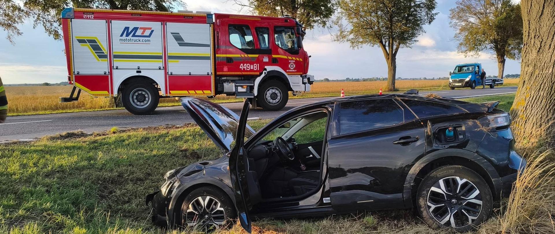 Pojazd pożarniczy w tle, na 1 planie auto osobowe biorące udział w zdarzeniu drogowym. Jest częściowo rozbite, znajduje się w rowie. 