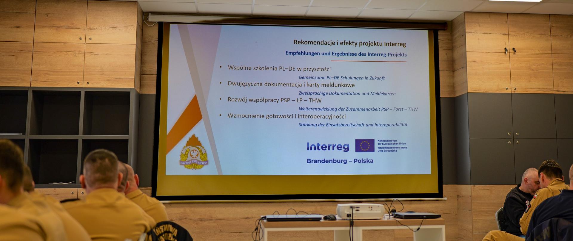 Na ekranie projekcyjnym widoczny slajd z napisem „Rekomendacje i efekty projektu Interreg”. Na sali siedzą strażacy w beżowych mundurach PSP, słuchając prezentacji dotyczącej współpracy polsko-niemieckiej w ramach programu Interreg Brandenburgia – Polska.