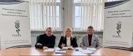 Trzy osoby siedzą przy stole konferencyjnym i przeglądają dokumenty. Kobieta pośrodku kartkuje teczkę, mężczyźni po bokach czytają pisma. Po prawej stronie widoczny roll-up Wojewódzkiego Inspektoratu Ochrony Roślin i Nasiennictwa w Rzeszowie, w tle okna z żaluzjami.