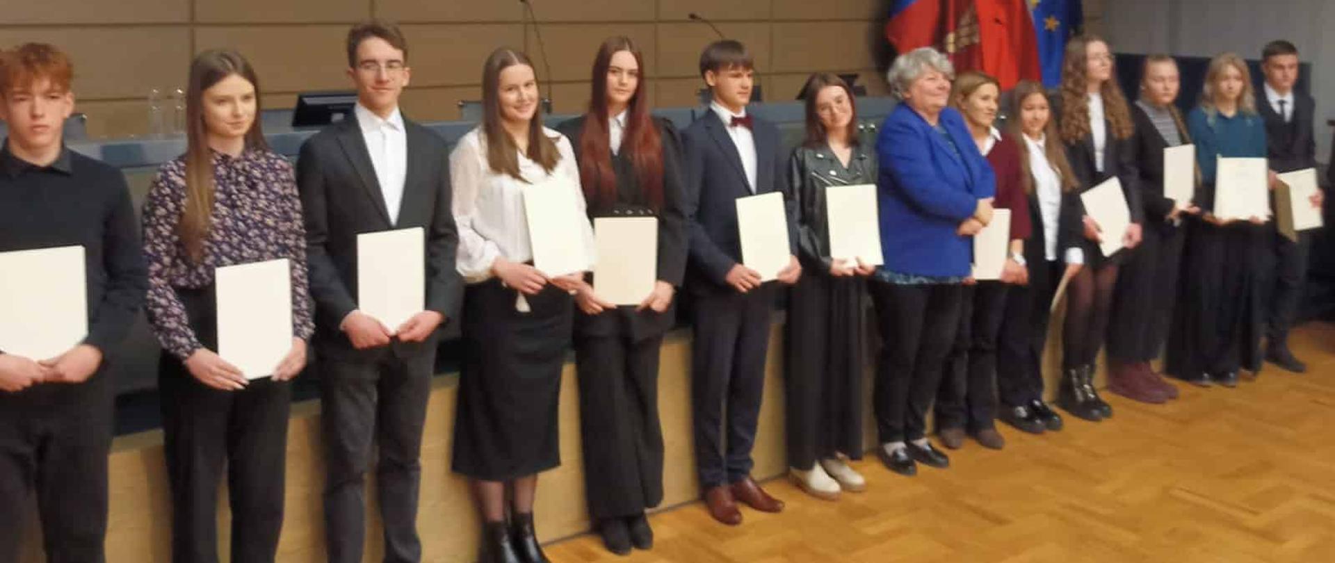 Pamiątkowe zdjęcie większej grupy stypendystów i przedstawicieli władz oświatowych stojących w rzędzie na scenie z dyplomami w dłoniach.
