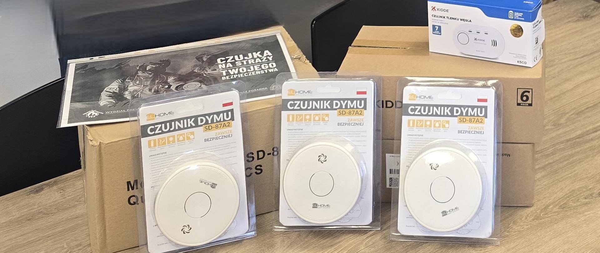 Zbliżenie na zapakowane czujniki dymu ustawione na stole oraz kartony z urządzeniami. Na jednym z kartonów leży broszura informacyjna.