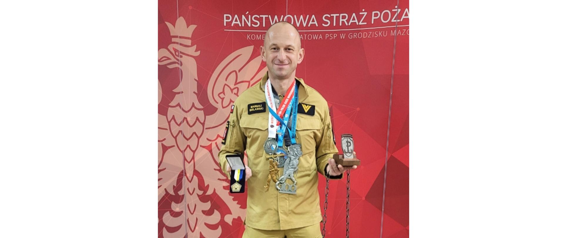 Pełne ujęcie stojącego strażaka w mundurze w kolorze khaki, z wieloma medalami zawieszonymi na szyi oraz trzymającego statuetkę i dużą obręcz z łańcuchem w dłoniach. W tle czerwona ściana z napisem "PAŃSTWOWA STRAŻ POŻARNA".