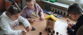 po obu stronach stołu siedzą dzieci i budują domek dla owadów. na stole leżą szyszki i rurki, dzieci trzymają w rękach nożyczki, pośrodku stoi nieukończony domek