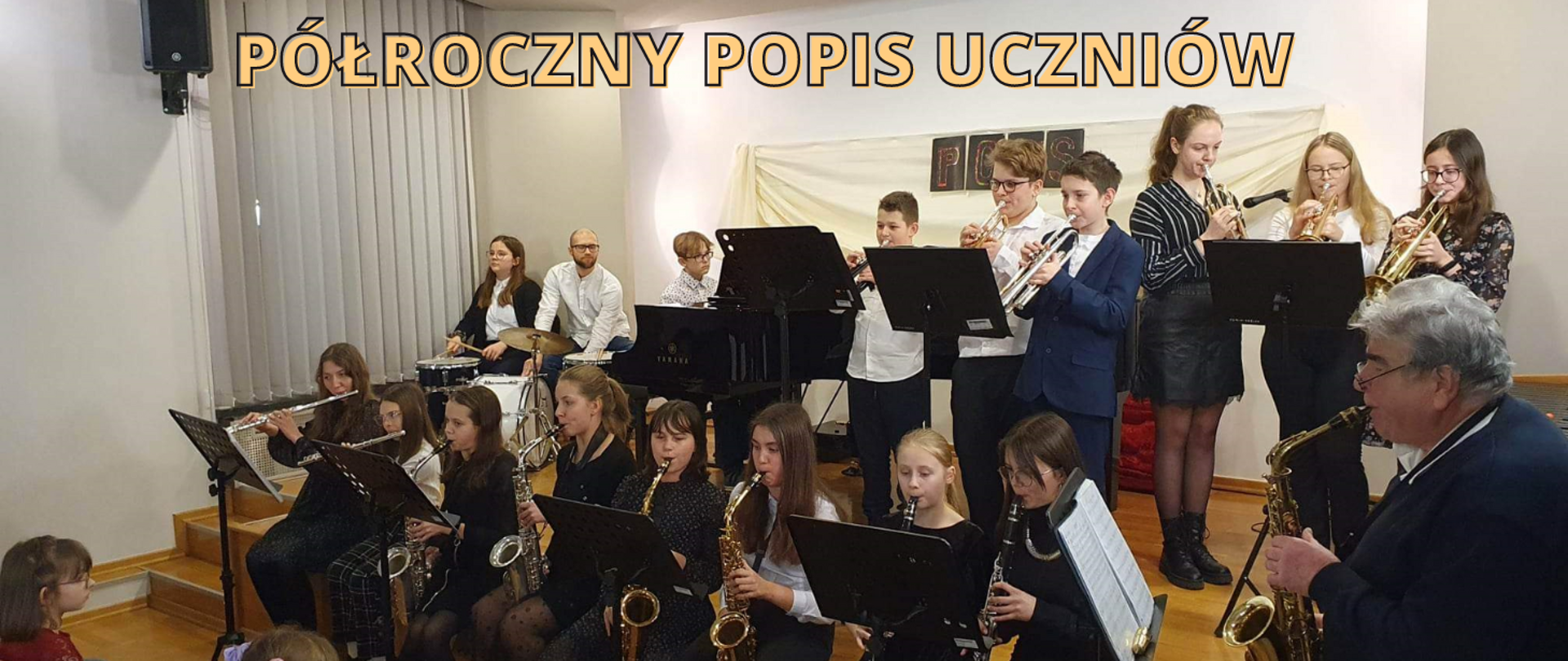 Na zdjęciu grupa ludzi. Wszyscy grają na instrumentach. Jedni na trąbce, inni na saksofonie, jeszcze inni na klarnecie, flecie poprzecznym. Dwie osoby grają na perkusji, a jeden chłopiec na fortepianie. Przed nimi stoją pulpity. Wszyscy razem tworzą szkolną orkiestrę dętą.