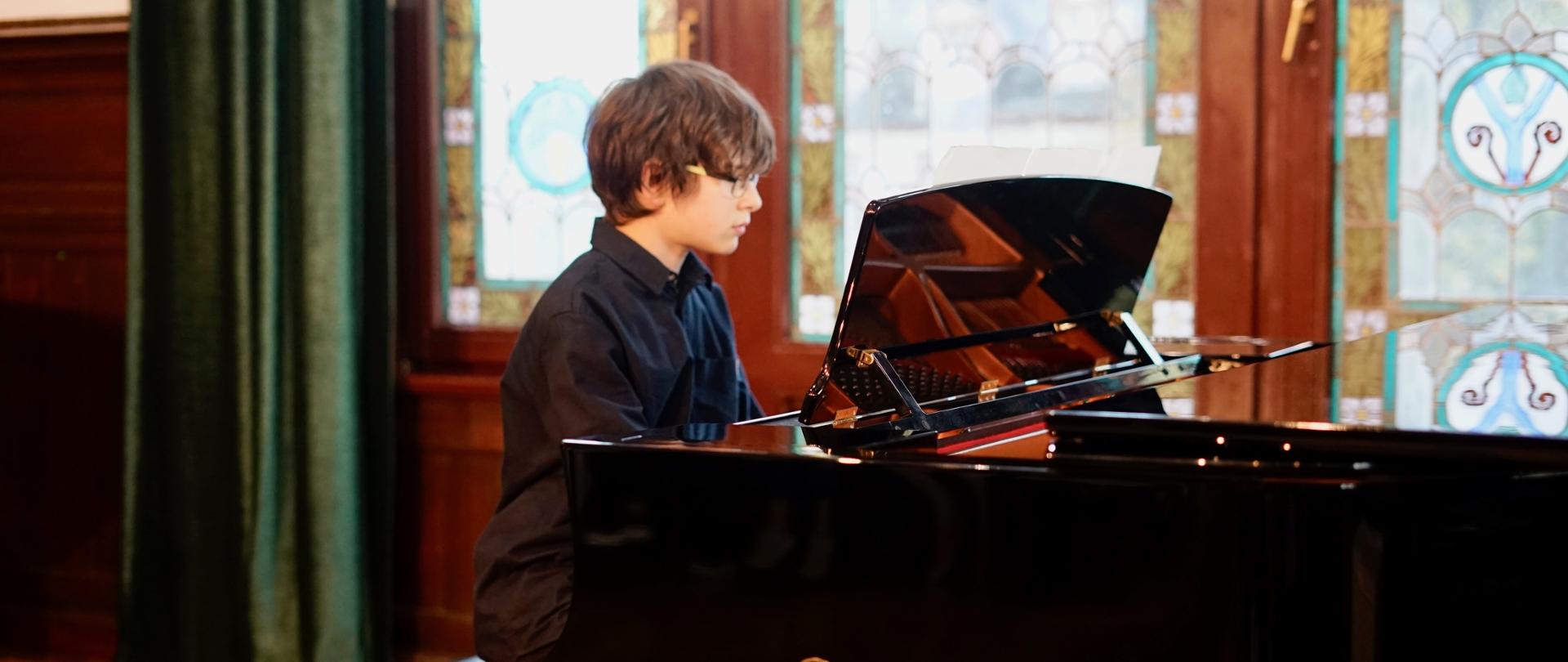 Zdjęcie przedstawia: od lewej fragment brązowej balustrady oraz ciemnozielonej kotary, w lewym dolnym rogu fragment jasnego parkietu. Na środku nastoletni chłopiec, o czarnych włosach w czarnej koszuli i okularach, siedzący przy czarnym fortepianie, ze złotym logiem i napisem yamaha, prawym bokiem skierowany do publiczności. W tle brązowe trzyskrzydłowe drzwi balkonowe, w oknach drzwi kolorowe witraże.