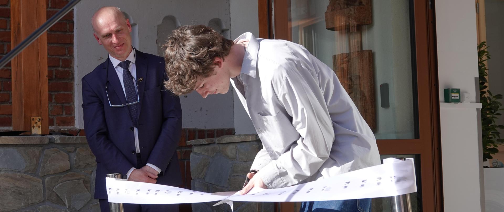 Na zdjęciu widzimy moment przecięcia symbolicznej wstęgi. Po lewej stronie widzimy Marka Króla-Józagę dyrektora szkoły ubranego w granatowy garnitur, biała koszulę z granatowym krawatem. Po prawej stronie widzimy Franciszka Wierciocha ucznia kl. 3A, Przewodniczącego Samorządu Uczniowskiego w PLSP im. A. Kenara w Zakopanem. Uczeń jest ubrany jasno-beżową koszulę i niebieskie spodnie. Przed postaciami na pierwszym planie rozwieszona jest na dwóch metalowych słupkach biała wstęga z nadrukowanymi czarnymi pegazami. Uczeń przecina wstęgę, co jest równoznaczne z otwarciem budynku szkoły po termomodernizacji. W tle widzimy główne wejście do budynku szkoły.