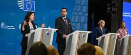 Na sali za kilkoma mównicami stoją cztery osoby, pierwsza z lewej kobieta w czarnej marynarce mówi do mikrofonu, obok niej stoi wiceminister Szeptycki, za nimi wielki niebieski ekran z logiem UE.