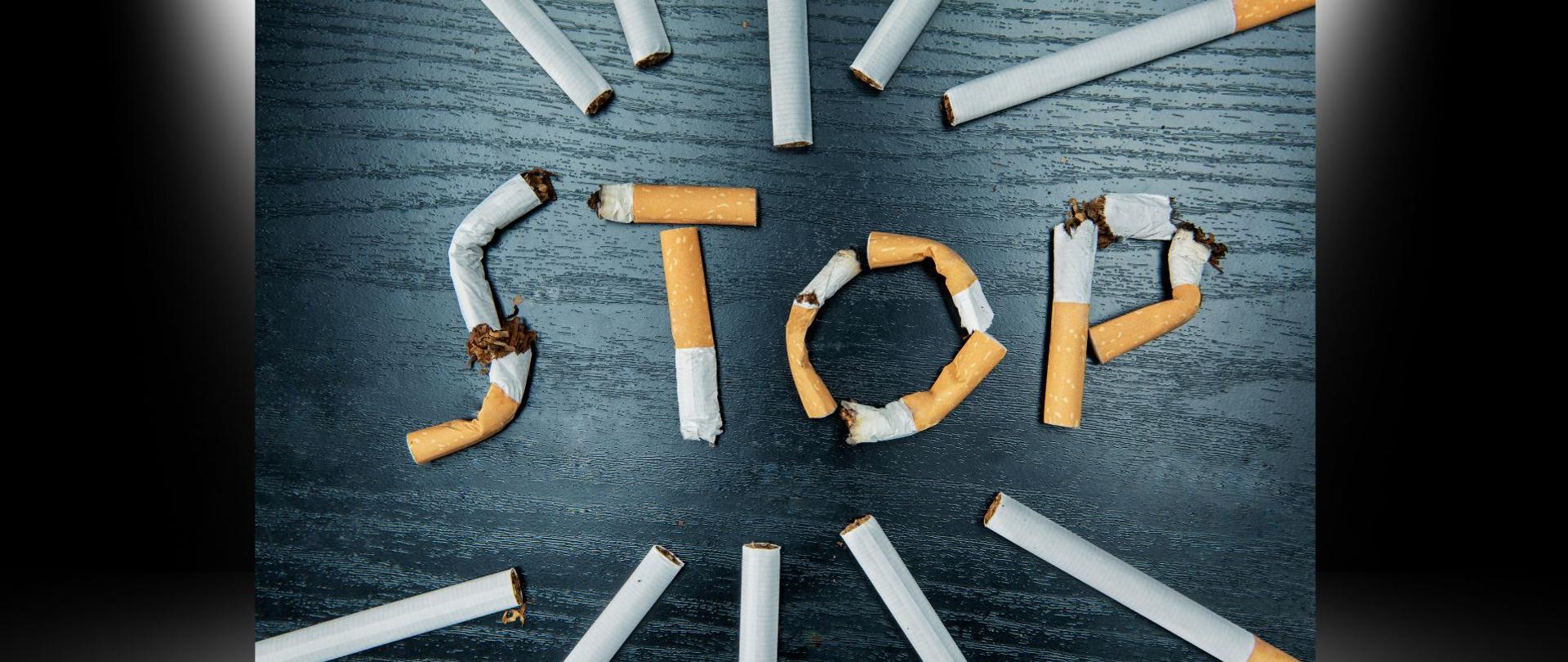 Kompozycja wizualna na ciemnym, drewnianym tle, w której niedopałki i połamane papierosy tworzą słowo STOP w centrum. Dookoła niego promieniście ułożono całe papierosy.