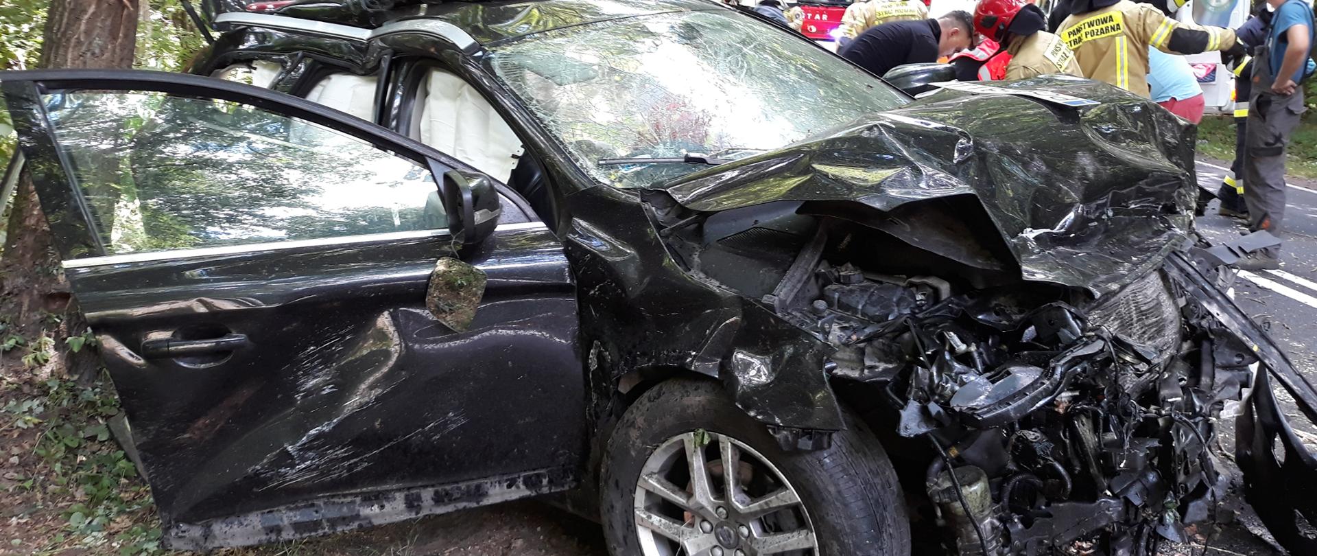 Słonecznie. Czarne auto osobowe kompletnie zniszczone po wypadku. W tle biała karetka ratownictwa medycznego i strażacy w paskowych mundurach i czerwonych hełmach oraz drzewa.