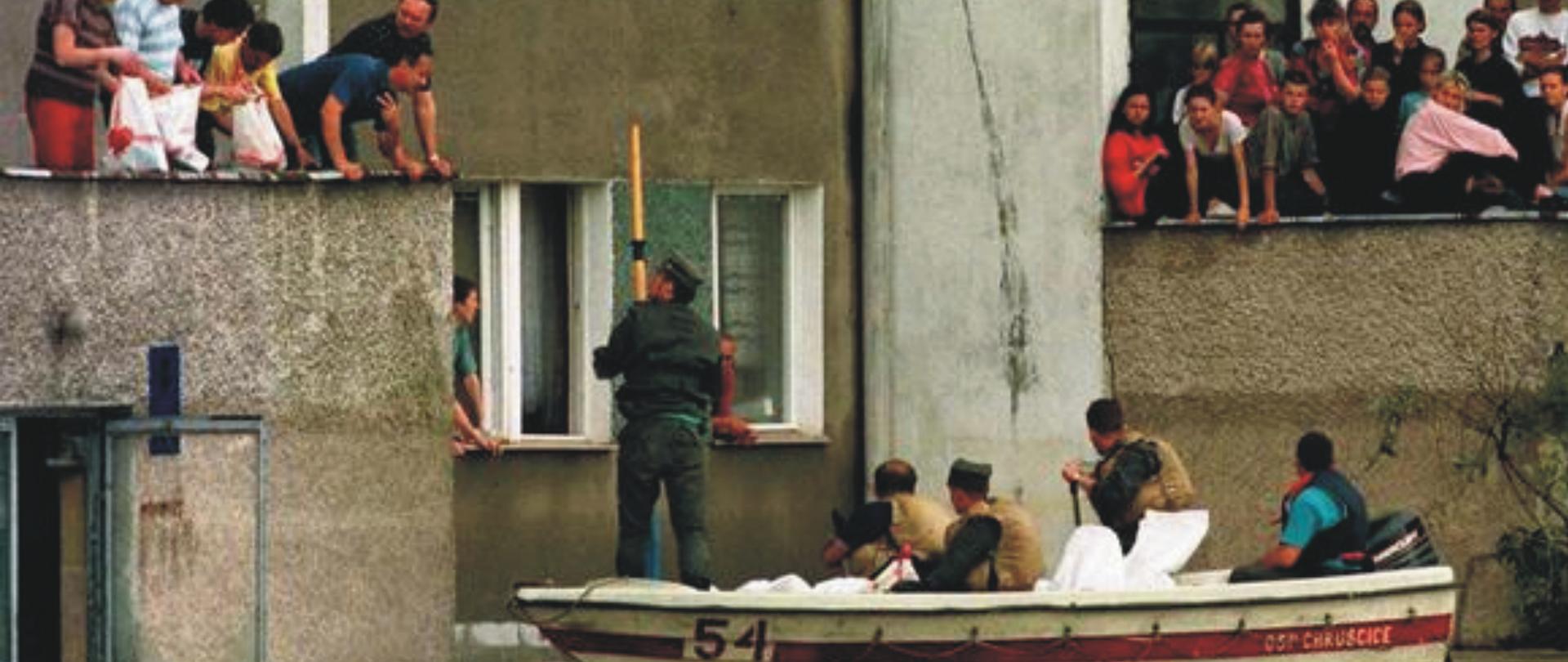 Na zdjęciu widać jak strażacy w łódce dostarczają żywność do mieszkańców przebywających w zalanych domach