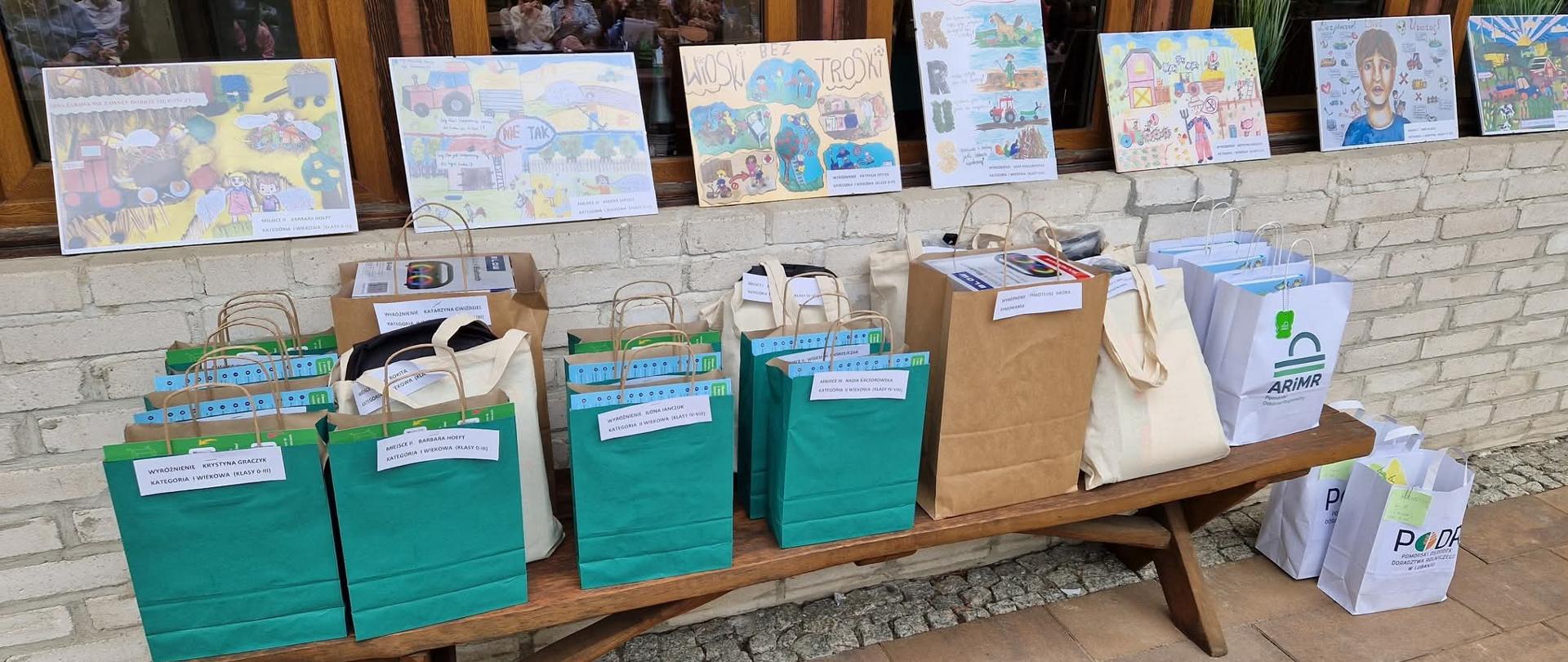 Na zewnątrz budynku na kamiennym obmurowaniu budynku wystawiono prace plastyczne dzieci. poniżej na ławce stoją kolorowe torby z logotypami organizatorów konkursu z nagrodami i naklejonymi karteczkami z nazwiskami laureatów.