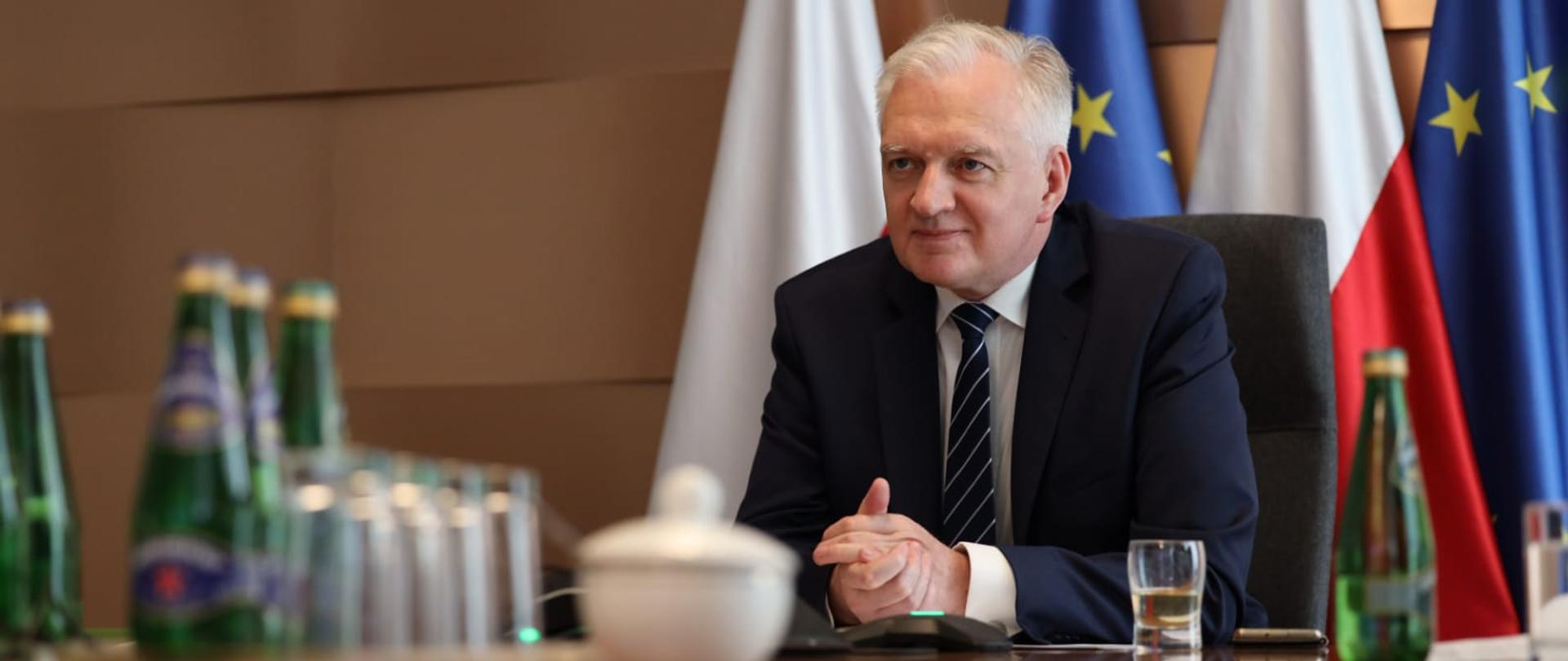 Wicepremier Jarosław Gowin podczas wideokonferencji. Siedzi za stołem na tle flag Polski i Unii Europejskiej.