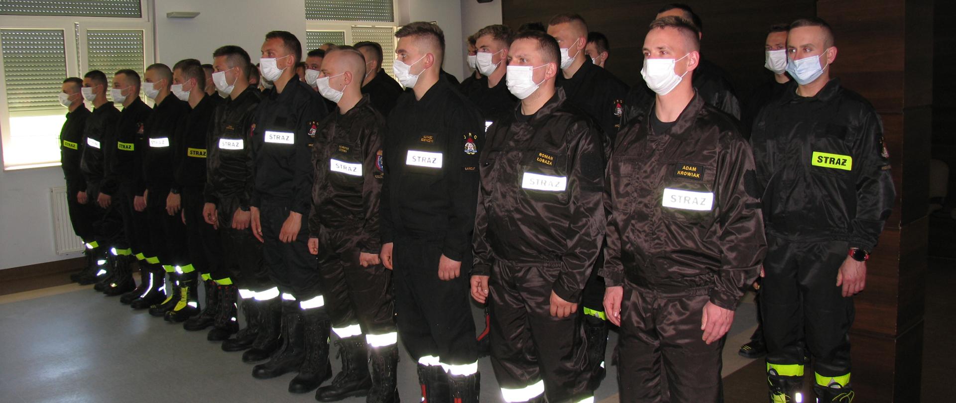 Na sali wykładowej są ustawienie strażacy w trzech szeregach z maseczkami na twarzy.
