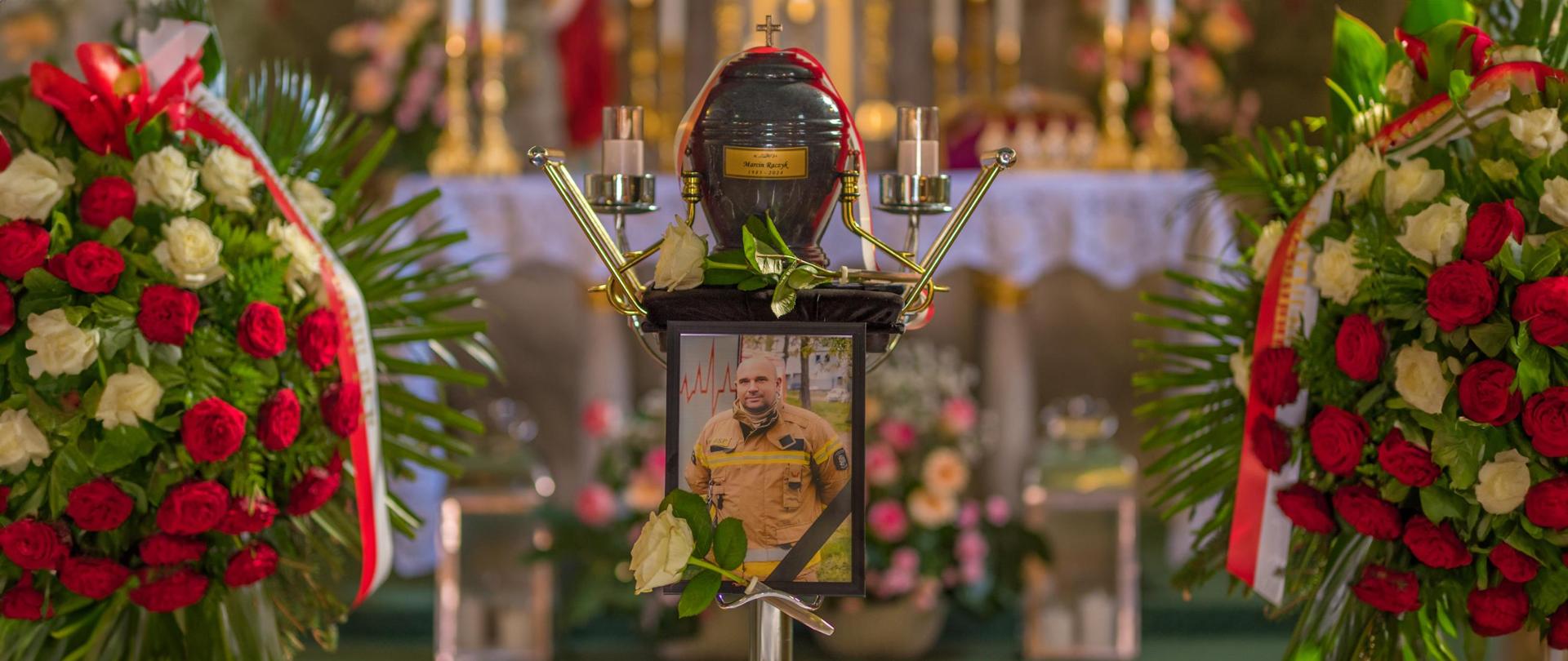 Urna z prochami zmarłego, pod urną zdjęcie strażaka w mundurze, po bokach wieńce z kwiatów