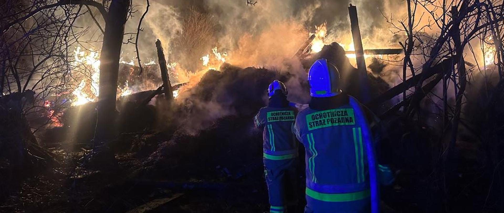 Na zdjęciu widać ogień wydostający się spod zawalonej stodoły. Widać także dwóch strażaków w umundurowaniu specjalnych, którzy trzymają w rękach wąż strażacki.