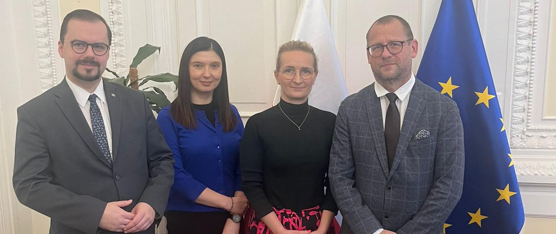 Dwóch mężczyzn w garniturach na zewnątrz, dwie kobiety elegancko ubrane stoją pośrodku