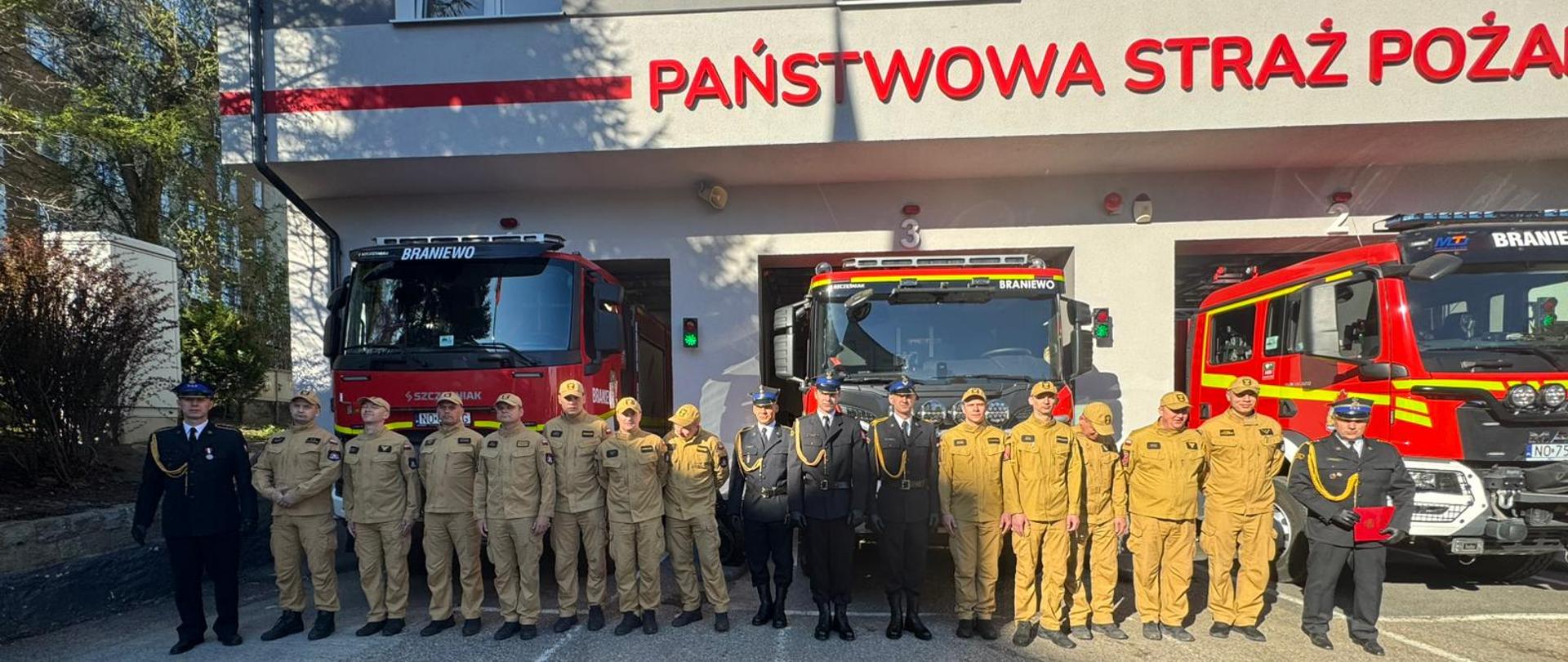 W jednym szeregu ustawieni strażacy na tle samochodów strażackich i remizy. Słonecznie.