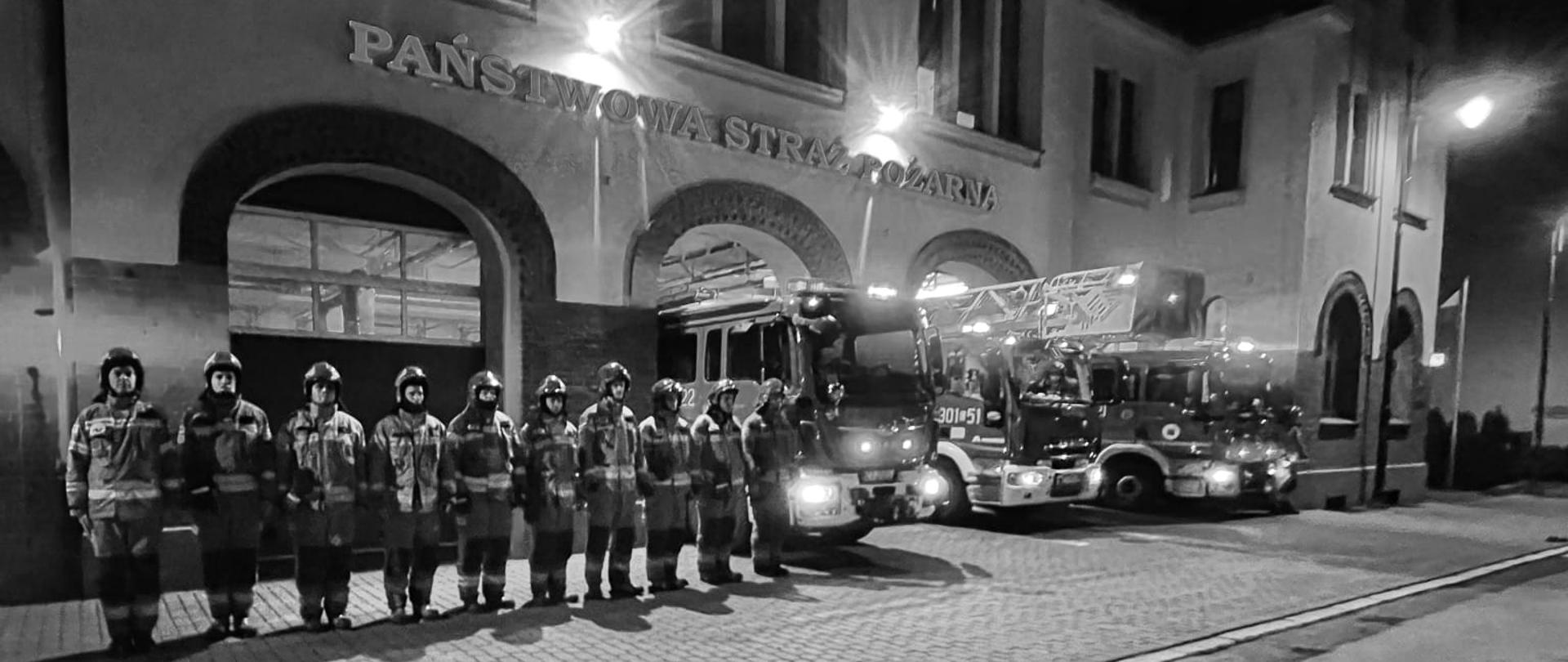 Strażacy na zbiórce w szeregu w ubraniach specjalnych, w tle przody wozów pożarniczych i frontowa elewacja budynku JRG1. Zdjęcie czarno-białe.