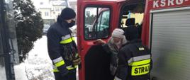 Zdjęcie zrobione w zimowej scenerii. Na zdjęciu widać dwóch strażaków ubranych w ubrania bojowe pomaga starszej kobiecie wysiąść ze strażackiego samochodu. Wszyscy mają maseczki na twarzach zasłaniające usta i nos.
