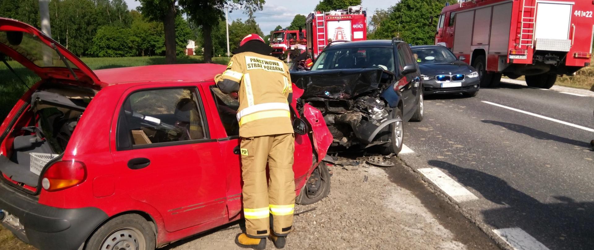 Zdjęcie przedstawia strażaka w ubraniu bojowym w trakcie prowadzenia działań przy czerwonym samochodzie Daewoo Matiz stojącym na poboczu drogi z uszkodzonym przodem naprzeciw pojazdu czarny SUV BMW z uszkodzonym przodem i komorą silnika na drodze ustawione samochody gaśnicze i cywilny pojazd Policji.