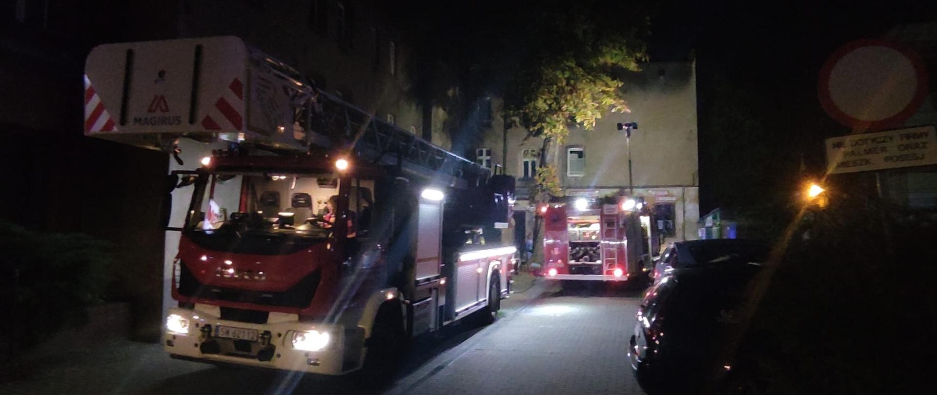 Na pierwszym planie drabina straży pożarnej stojąca na ulicy, w tle stoi samochód gaśniczy stojący przy budynku objętym pożarem.