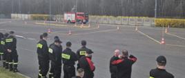 Na zdjęciu strażacy stojący przy placu manewrowym, na którym jeden z kierowców w samochodzie ratowniczo-gaśniczym wjeżdża w wyznaczone pachołkami ciasne miejsca tak aby nie potrącić żadnego z nich.