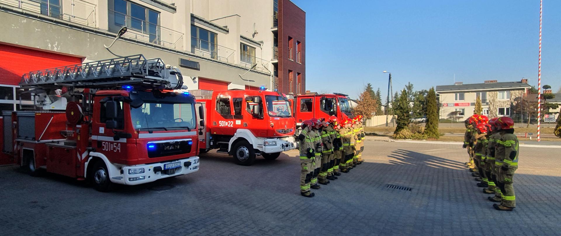 Po prawej stronie zdjęcia strażacy ustawieni w dwóch rzędach po prawej i lewej stronie podczas uroczystej zmiany służby, stojący na baczność, pierwszy z lewej i ostatni z prawej oddają honory. Z lewej strony trzy samochody pożarnicze na tle budynku komendy.