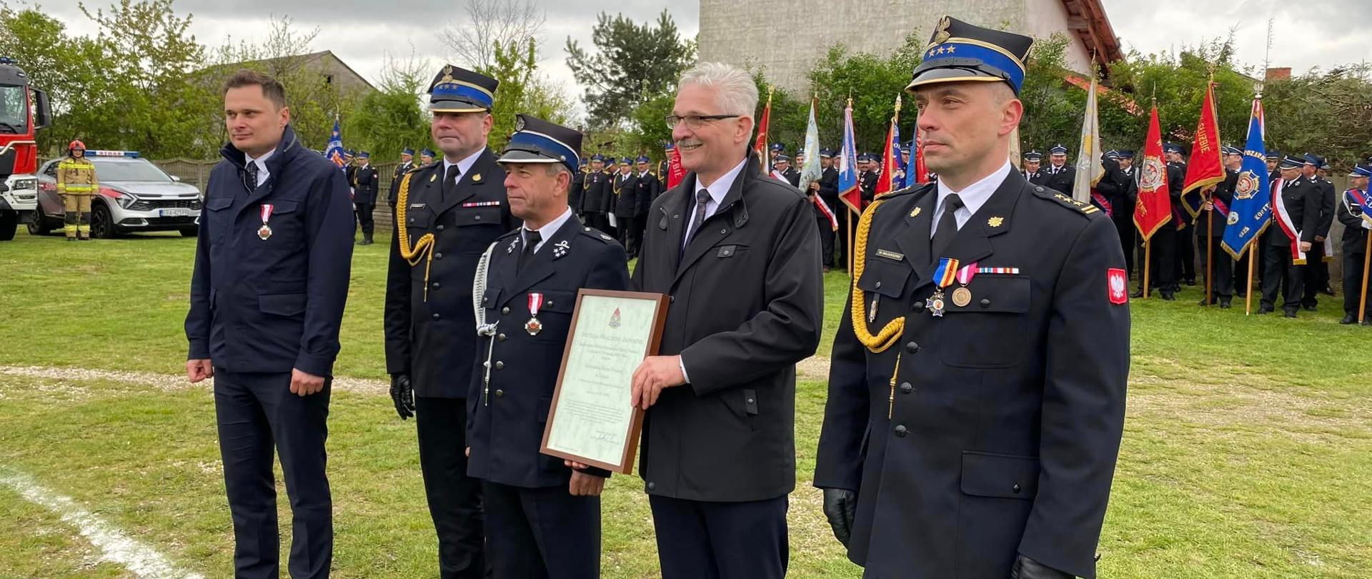 od lewej minister Ciecióra, st. bryg. Sebastian Kościsty, prezes osp Żytno, wójt Gminy i komendant powiatowy PSP w Radomsku, wójt gminy prezentuje akt włączenia jednostki do KSRG, w tle strażacy i poczty sztandarowe