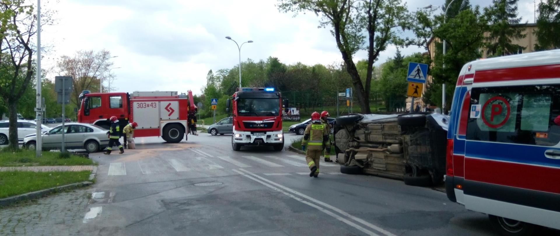Zdjęcie przedstawia przewrócony na bok samochód marki Nissan. Z prawej strony znajduje się karetka Pogotowia Ratunkowego natomiast na wprost znajdują się dwa samochody strażackie oraz strażacy stojący przy przewróconym aucie.