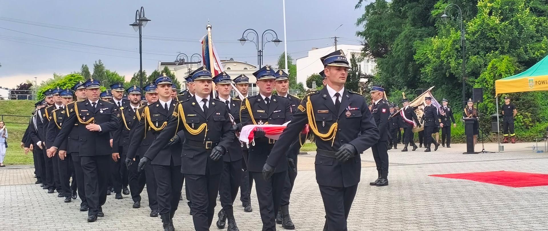 Uroczysty marsz strażaków w galowych mundurach Państwowej Straży Pożarnej. Na pierwszym planie widać idących ramię w ramię funkcjonariuszy w ciemnogranatowych garniturach, czapkach rogatywkach i złotych sznurach galowych. Dwóch z nich niesie złożoną flagę Polski. W tle maszeruje kolumna kolejnych strażaków, a za nimi widoczne są zielone drzewa, maszty i latarnie na placu miejskim.