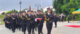 Uroczysty marsz strażaków w galowych mundurach Państwowej Straży Pożarnej. Na pierwszym planie widać idących ramię w ramię funkcjonariuszy w ciemnogranatowych garniturach, czapkach rogatywkach i złotych sznurach galowych. Dwóch z nich niesie złożoną flagę Polski. W tle maszeruje kolumna kolejnych strażaków, a za nimi widoczne są zielone drzewa, maszty i latarnie na placu miejskim.
