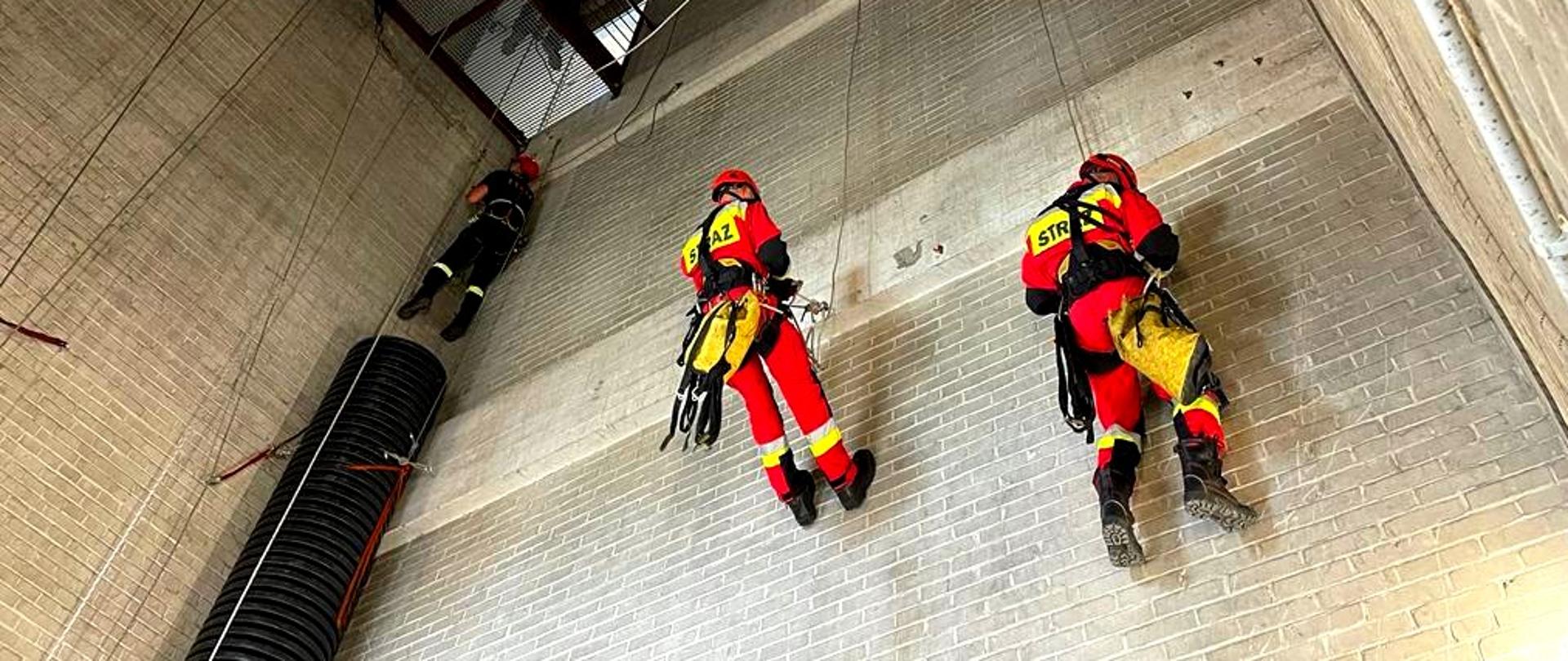 Dwóch strażaków w pomarańczowych kombinezonach wspina się na linach po ścianie