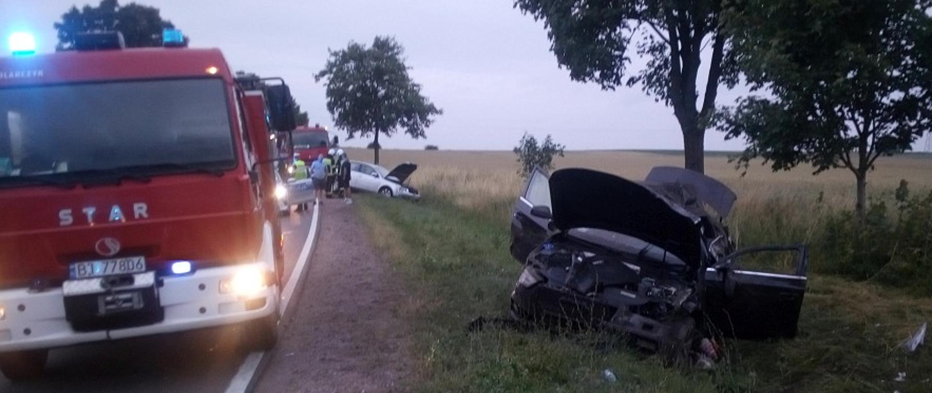 Na pierwszym planie widać rozbite auto osobowe znajdujące się w rowie, obok na jezdni stoi pojazd pożarniczy na sygnałach świetlnych, w tle znajduje sie drugie auto w rowie i samochód strażacki.