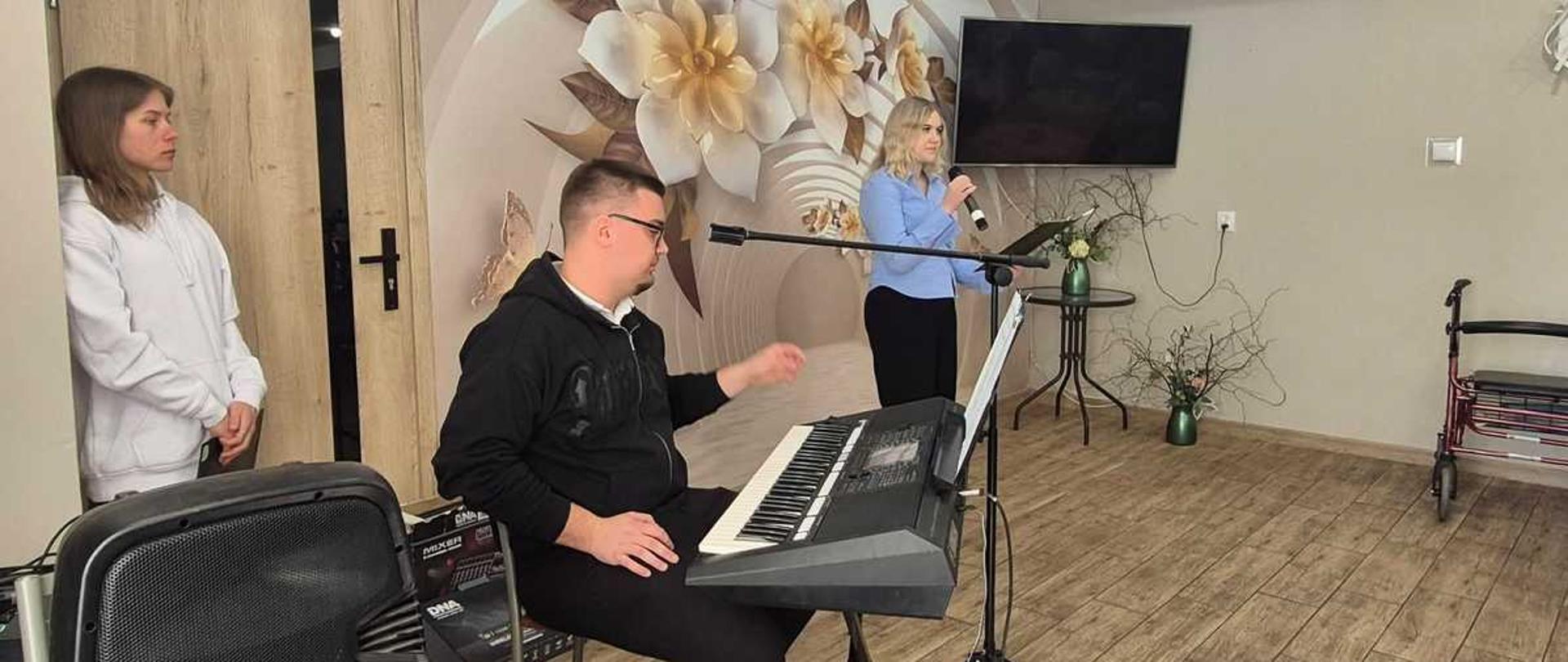 Główny plan
Pośrodku, nieco po lewej: Mężczyzna w okularach i czarnej bluzie z kapturem siedzi na krześle i gra na elektronicznym keyboardzie. Instrument spoczywa na czarnym, metalowym statywie.
W głębi, po prawej od muzyka: Kobieta o blond włosach, ubrana w jasnoniebieską koszulę i czarne spodnie, śpiewa do mikrofonu. Przed nią, na statywie, znajduje się nutnik z tekstami piosenek.
Po lewej stronie: Młoda dziewczyna w białej bluzie z kapturem stoi spokojnie przy drzwiach, obserwując występ.
Otoczenie i detale
Tło: Ścianę za muzykami zdobi duża fototapeta z motywem jasnych kwiatów. Obok wisi wyłączony telewizor.
Wyposażenie: Na podłodze imitującej drewniane panele stoi duża czarna kolumna głośnikowa (na pierwszym planie po lewej) oraz chodzik rehabilitacyjny (pod ścianą po prawej).
Wystrój: W pokoju widać również mały stolik z wazonem z kwiatami oraz delikatne ozdoby roślinne na ścianach. Oświetlenie jest jasne i punktowe, umieszczone w podwieszanym suficie.
Atmosfera zdjęcia wydaje się spokojna i skupiona, sugerując występ w kameralnym gronie, np. w domu opieki lub świetlicy.