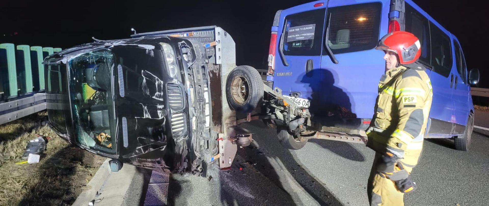 Na zdjęciu znajduje się strażak ubrany w ubranie specjalne, w tle widać dwa wraki pojazdów