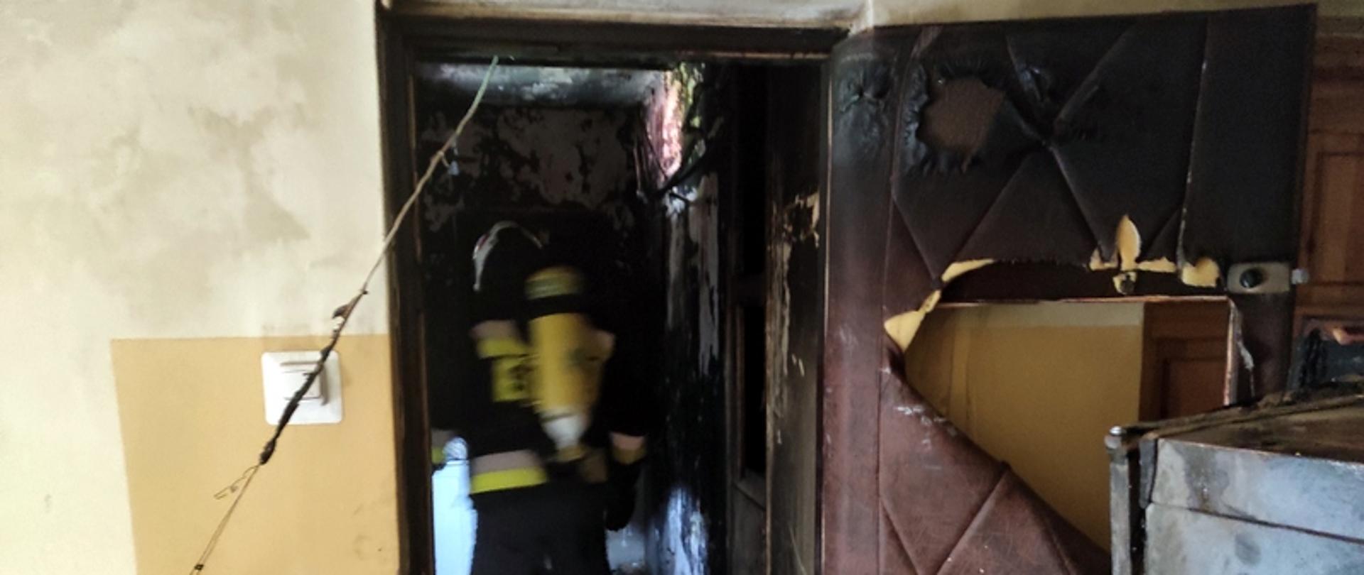 na zdjęciu widać otwarte drzwi do mieszkania, w którym doszło do pożaru. Widać sylwetkę strażaka w mundurze bojowym wewnątrz.