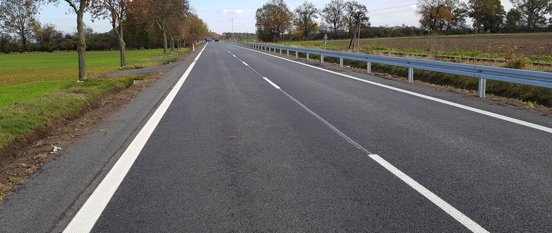 Zdjęcie przedstawia drogę krajową. Po jednym pasie w każdym kierunku. Z prawej strony drogi widoczna bariera ochronna. W oddali widoczne samochody. 