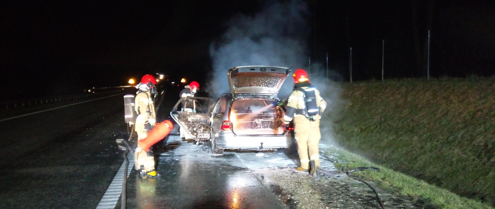 Zdjęcie przedstawia samochód mercedes z otwartą klapą bagażnika z którego wydobywa się biały dym. Obok samochodu znajdują się strażacy którzy gaszą pożar. Jest noc. Samochód znajduje się na poboczu drogi.